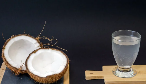 Fruta exótica de coco, dos mitades de la fruta en la cáscara, fondo oscuro, un vaso de agua de coco en la cocina y pizarra, enfoque selectivo —  Fotos de Stock