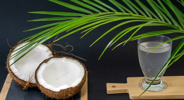 Fruta exótica Coco, dos mitades de fruta en la cáscara, fondo oscuro, un vaso de agua de coco en la cocina y pizarra, palmeras verdes, enfoque selectivo —  Fotos de Stock