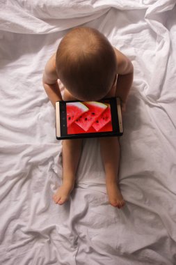 Küçük bir çocuk beyaz bir levha bir tablet ile oynarken bir yatak üzerinde oturuyor