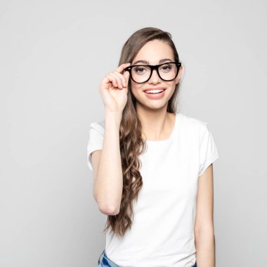 Kameraya gülümseyen beyaz tişört ve inek gözlük, dostu uzun saçlı kız öğrenci portresi. Gri arka planı vurdu stüdyo.