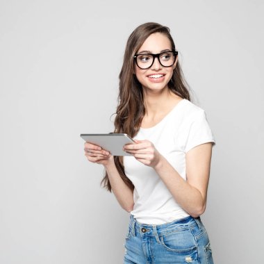 Portrait of beautiful long hair female student wearing white t-shirt and nerd glasses, holding a digital tablet. Studio shot against grey background. clipart