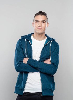 Studio shot of cool young man wearing hooded shirt standing with arms crossed against grey background, smiling at camera.  clipart