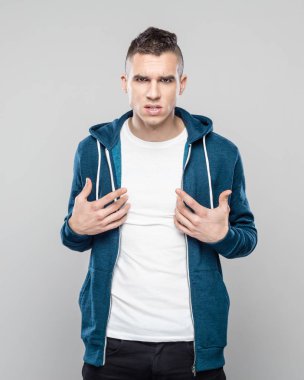 Kızgın genç adam gri arka plan üzerinde duran ve kameraya bakarak onun beyaz t-shirt işaret kukuletalı gömlek giymiş stüdyo çekim. 