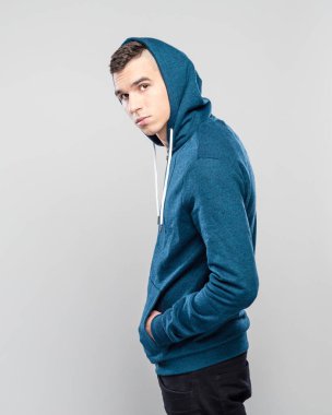 Studio shot of cool young man wearing hooded shirt standing against grey background with hands in pockets, looking at camera. clipart