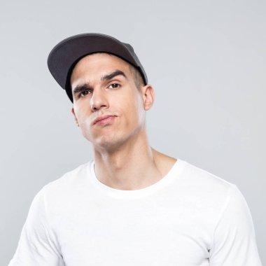 Stüdyo memnun genç adamın beyaz t-shirt ve gri arka plan üzerinde duran beyzbol şapkası giyen kameraya seyir. Yüzünü kapat.