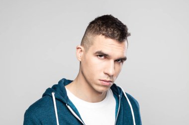 Kızgın genç adam gri bir arka plana dayanır kameraya bakarak, ayakta kukuletalı gömlek giymiş, stüdyo vurdu. Yüzünü kapat.