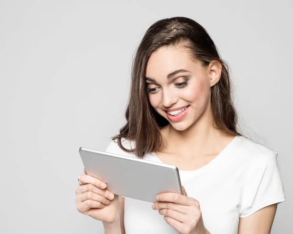 Dijital tablet kullanma beyaz t-shirt giyen heyecanlı genç kadın portresi. Gri arka planı vurdu stüdyo.