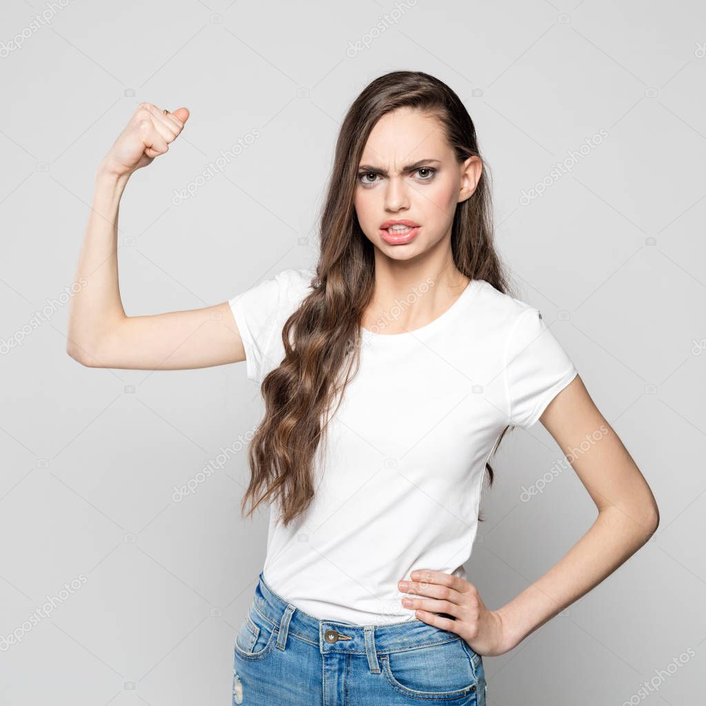 Studio portrait of angry young woman wearing white t-shirt and jeans flexing her muscles, staring at camera. Grey background.