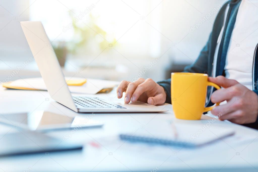 Indoors shot of man sitting by the desk and typing on laptop, close up of hands, unrecognizable person.
