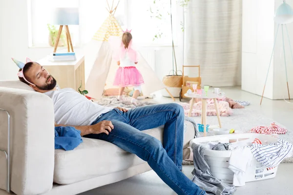 Young single father overwhelmed with household chores sleeping on sofa in messy living room with his little daughter playing in the background.