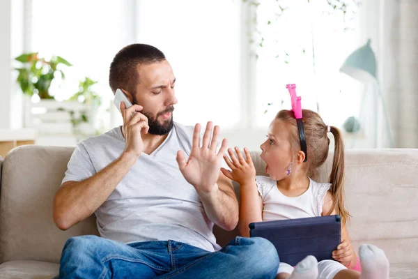Oturma odasında kanepede oturan ve onu rahatsız litte kızı süre telefonda konuşurken genç baba. 