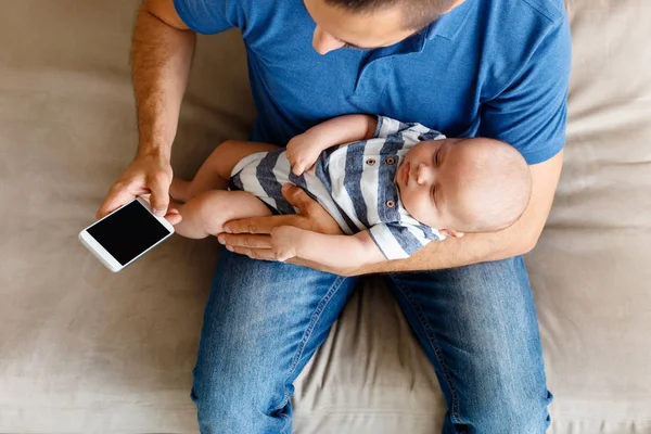 Bir evde koltukta oturan, bebek evlat holding ve akıllı telefon kullanan genç baba üstten görünüm. 