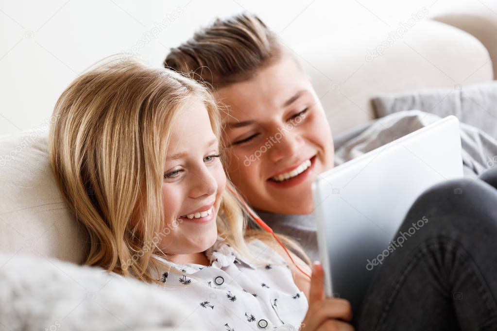 Happy brother and sister sitting on sofa at home in the living room and using digital tablet together.