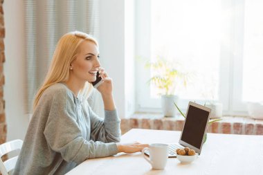 Masada evde oturan, kullanarak dizüstü bilgisayar ve cep telefonu üzerinde konuşurken güzel genç kadın yan görünüm. 