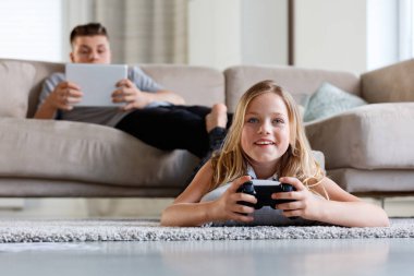 Kız ve erkek kardeşim evde zaman harcama. Yere yat döşeme ve kanepede oturan ve dijital tablet kullanarak çocuk video oyunu oynayan kız. 