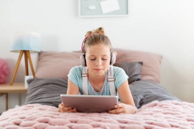 Yatak odasında yüzüstü yatarken ve dijital tablet kullanma kulaklık giyen kız. 