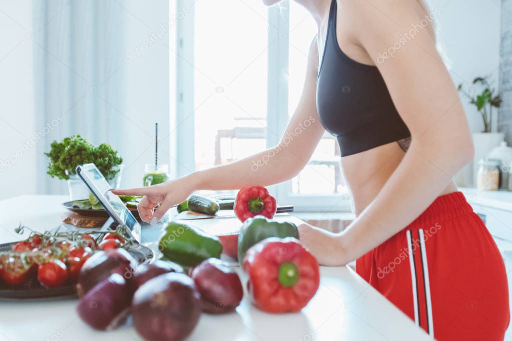 Young woman wearing sport clothes standing in the kitchen and preparing vegetarian food, using a digital tablet. Unrecognizable person. 