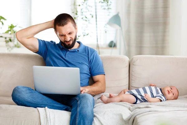 Worried young father working from home, using laptop while his baby boy lying on sofa next to him.