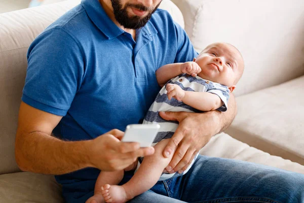 Close up of young father sitting on sofa at home, holding baby son and using smart phone.