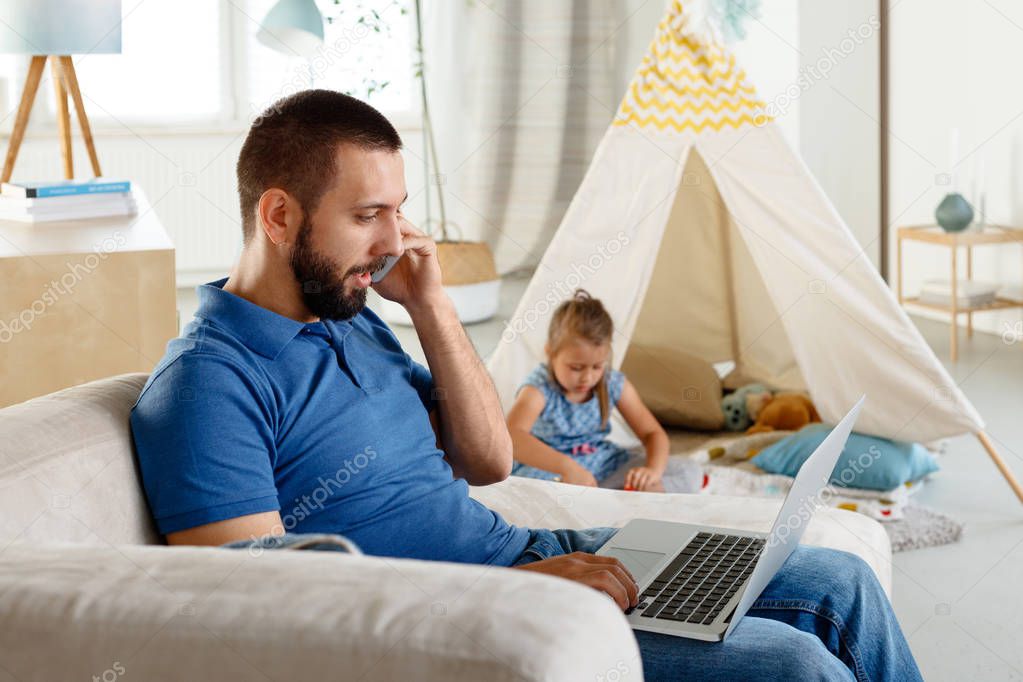 Young man sitting on sofa at home, talking on phone and using laptop while his daughter playing in the background. 