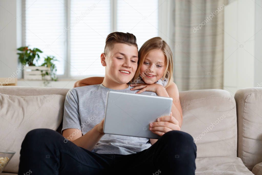 Brother and sister sitting on sofa at home in the living room and using digital tablet together.