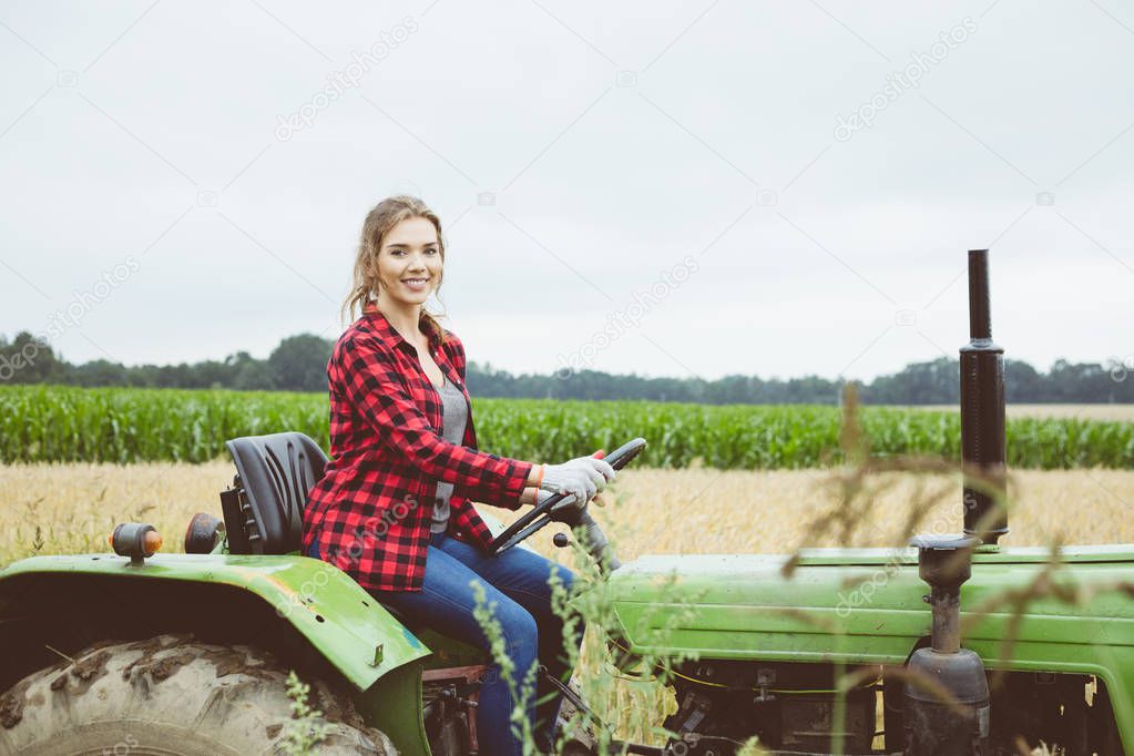 Outdoor shot of happy young woman sitting on tractor with corn filed in the background.