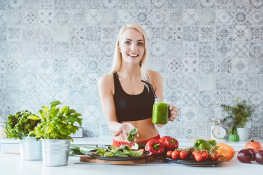 İçki ve ıspanak elinde, kameraya gülümseyen tutan hazırlanıyor yeşil smoothie mutfakta, Sarışın genç kadın spor giyim elbise. 