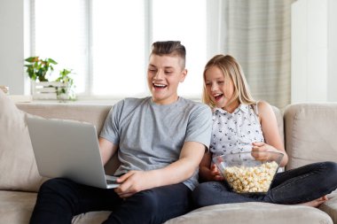 Teenage boy and his sister sitting on sofa at home and watching funny movie on laptop, eating popcorn.  clipart
