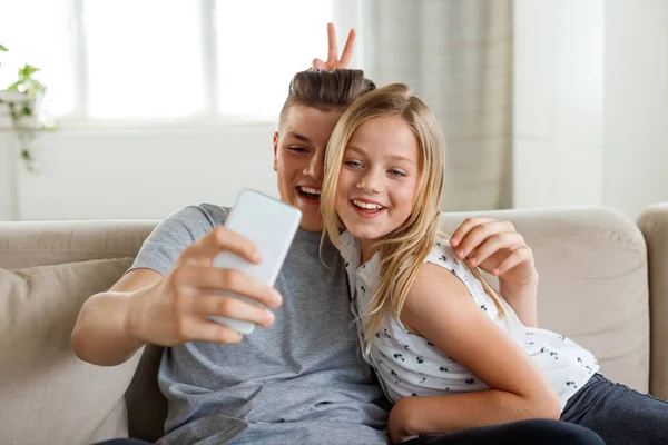 Happy brother and sister sitting on sofa in living room and taking selfie using smart phone.