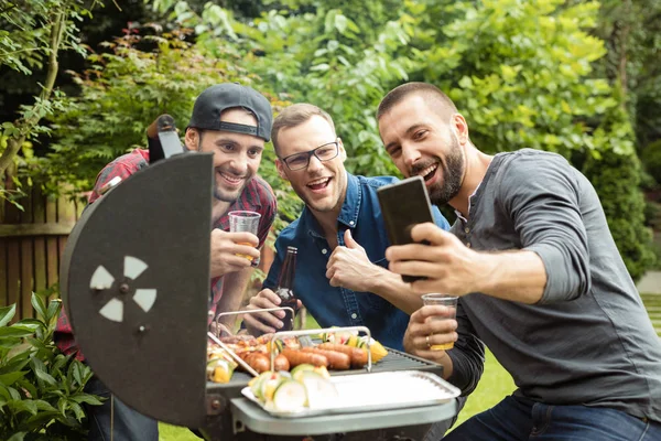 Three happy man enjoying outdoor party, standing next to barbecue grill and taking selfie.