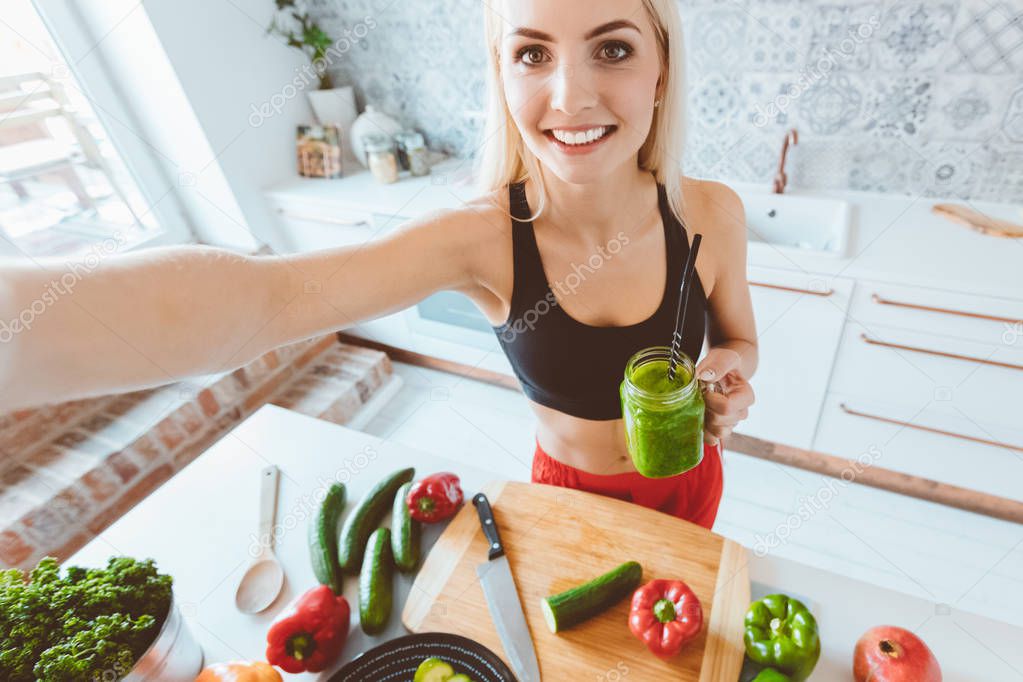 Indoor shot of young woman standing in the kitchen, holding green smoothie in hand and taking selfie. 