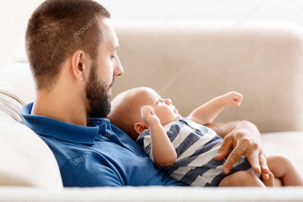 Happy young father sitting on sofa ant home and embracing his baby boy. 