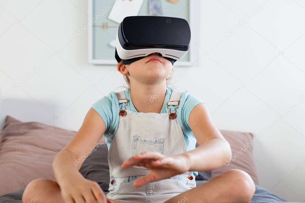 Cute girl sitting on bed in her bedroom and using virtual reality goggle. 