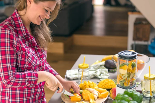 Young smiling woman making smoothie with fresh greens in the blender in kitchen at home.