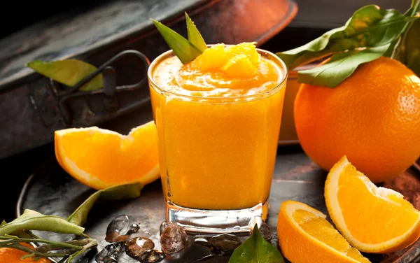 Orange smoothie and orange fruits with green leaves on dark background