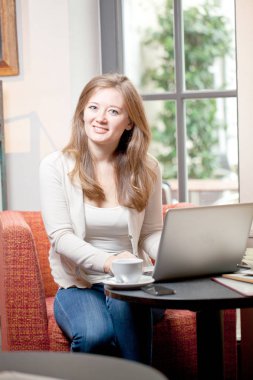 Ahşap masa kahve dükkanında otururken, kahve içme ve smartphone kullanarak genç kadın. Masada dizüstü bilgisayardır. Kız sohbet, internet, bloglama tarama. Kadın telefon tutarak ve onun ekranda arıyor.