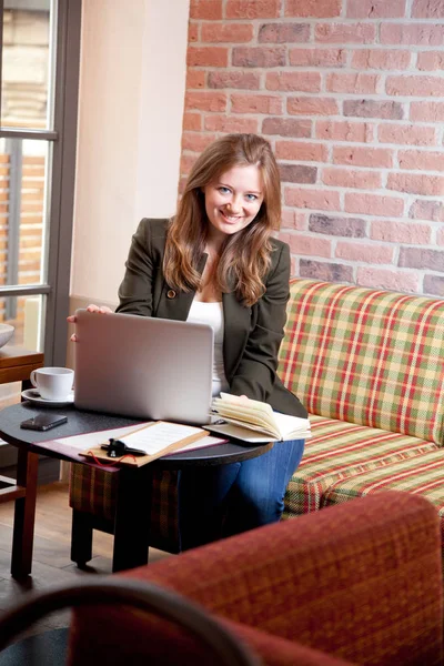 Ahşap masa kahve dükkanında otururken, kahve içme ve smartphone kullanarak genç kadın. Masada dizüstü bilgisayardır. Kız sohbet, internet, bloglama tarama. Kadın telefon tutarak ve onun ekranda arıyor.