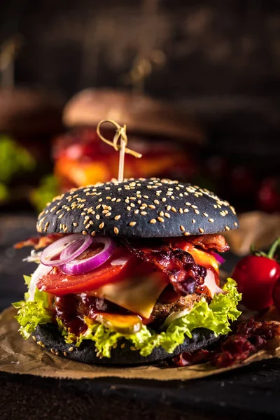 Black burger with meat patty, cheese, tomatoes, mayonnaise. Dark wooden rustic table. Modern fast food lunch.