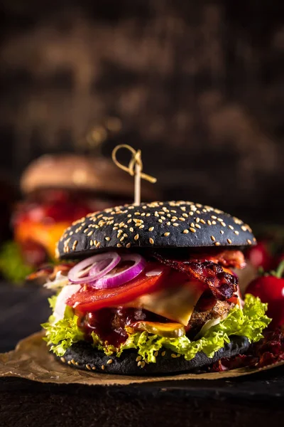 Black burger with meat patty, cheese, tomatoes, mayonnaise. Dark wooden rustic table. Modern fast food lunch.