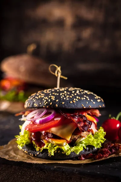 Black burger with meat patty, cheese, tomatoes, mayonnaise. Dark wooden rustic table. Modern fast food lunch.