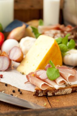 Lezzetli yemek, ekmek dilimlenmiş jambon, peynir, baharatlar ve sebzeler ahşap masada.