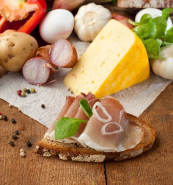 Lezzetli yemek, ekmek dilimlenmiş jambon, peynir, baharatlar ve sebzeler ahşap masada.