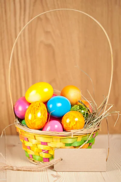 wicker basket with colorful painted easter eggs and blank label on wooden surface