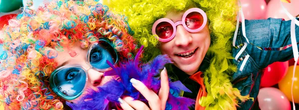 Funny young couple in colorful wigs and glasses posing against colorful party balloon background