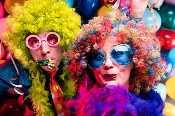 Funny young couple in colorful wigs and glasses having fun against colorful party balloon background