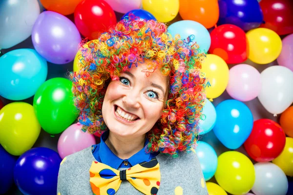 funny young woman in colorful wig posing against colorful party balloon background with dozens of balloons