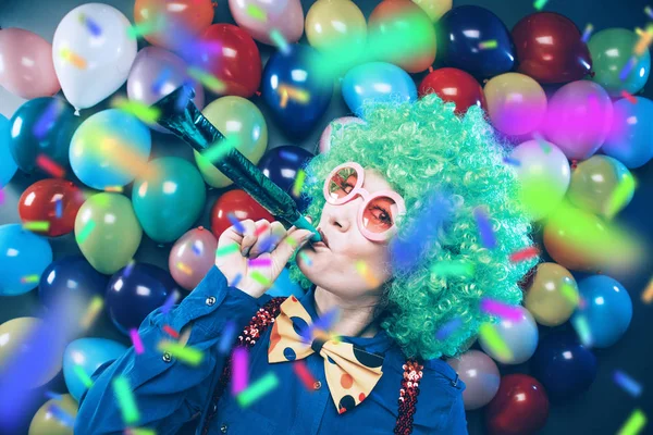 funny young woman in green wig and glasses posing against colorful party balloon background with dozens of balloons
