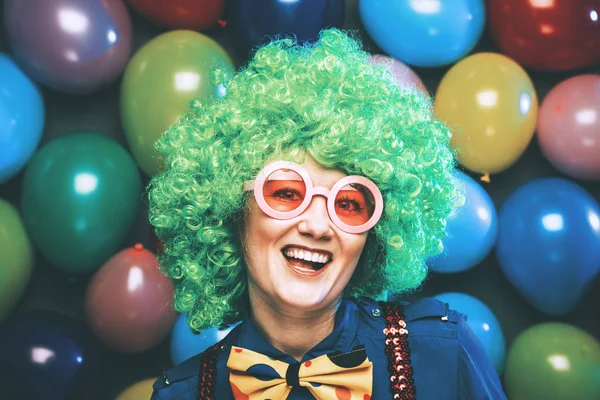 funny young woman in green wig and glasses posing against colorful party balloon background with dozens of balloons