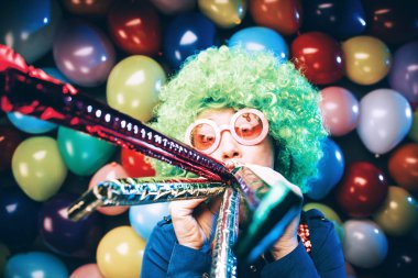 funny young woman in green wig and glasses posing against colorful party balloon background with dozens of balloons clipart