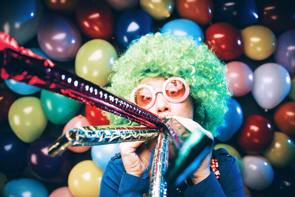 funny young woman in green wig and glasses posing against colorful party balloon background with dozens of balloons